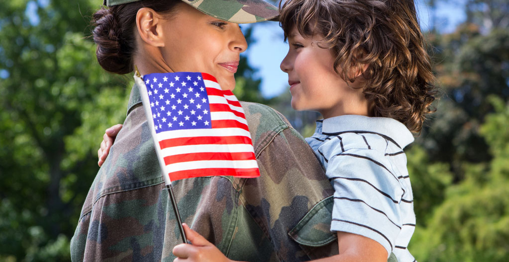 Young caucasian female veteran dressed in military fatigues holding a caucasian boy waving a small American flag