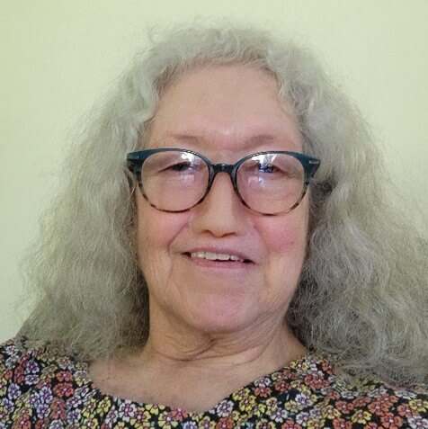 Photo of Barb, a woman with shoulder length gray hair and glasses. She is smiling at the camera and wearing black shirt 