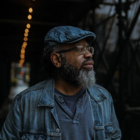 Photo of Dre, a man with short black hair under a blue cabbie cap, glasses and black and grey beard wearing blue shirt and denim