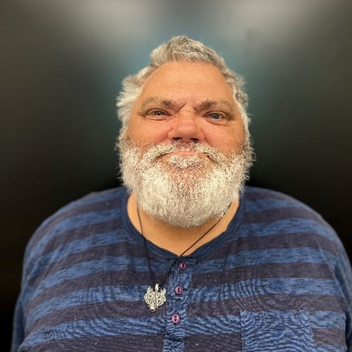 Photo of Eddie, a man with short grey hair, grey beard, and he is looking at the camera. He is wearing a black and blue striped pattern shirt. The background is a black wall.