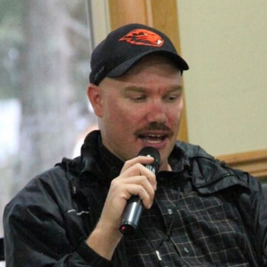 Photo of Justin, a man with a mustache, black ball cap  holding a microphone in his right hand