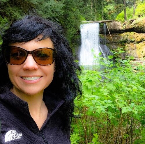 Photo of Krista, a woman with long black hair, dark sunglasses, wearing a zip up collar jacket in front of a nature scene