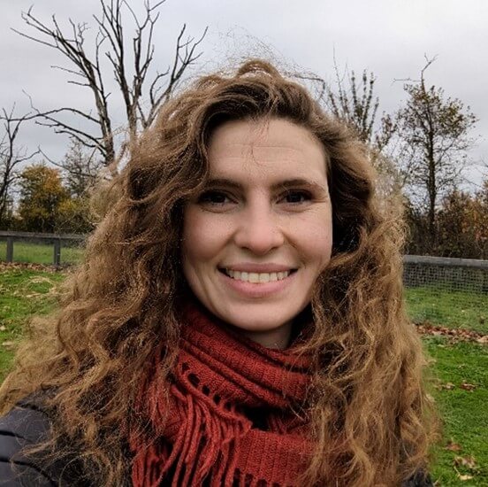 Photo of Rhiannon, a woman with long light brown curly hair wearing a black jacket with red scarf.